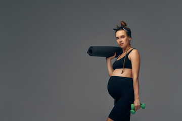 Fit young woman exercising during her pregnancy standing smiling at the camera with bottled water in one hand and a gym mat in the other