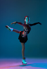 Dynamic portrait of young girl, female figure skater in black stage dress skating isolated on blue background in neon light. Concept of sport, beauty, active lifestyle.