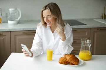A young smiling girl in a white shirt has breakfast with croissants with orange juice and uses a...