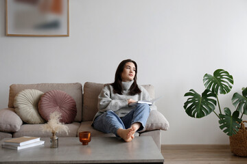 Young beautiful woman wearing grey turtleneck reading a book at home. Brunette female in a sweater sitting on beige textile couch in her lofty apartment. Background, copy space, close up.