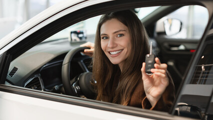 Fototapeta na wymiar Cheerful young woman holding key from auto, buying new car at showroom, sitting inside automobile salon at dealership