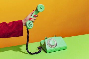 Colorful bright image of female hand holding old-fashioned green colored phone, picking up handset isolated over orange background
