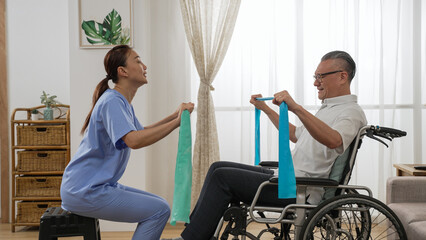 asian woman physiotherapist teaching handicapped older man how to use elastic band while she is doing resistance training on wheelchair at home
