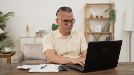 Fototapeta na wymiar Portrait of a nice asian grey hair man with glasses, working at home on some project, he is sitting at a white table looking at his laptop in front of him. Focus on the man