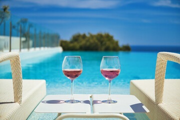 Two glasses with cocktails on table between deck chairs sunbeds on the swimming pool, sea and blue sky background. Summer holidays, relaxation, accommodation in luxury hotel concept, no people. Spain - obrazy, fototapety, plakaty