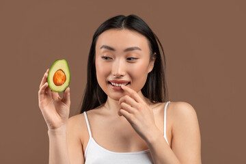 Natural skincare concept. Young asian woman holding ripe fresh avocado half, standing on brown background