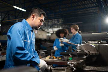 Selective focus of Asian male lathe worker in a blue uniform and gloves, standing working with cross feed handwheel of a lathe machine with blurred colleagues working in background in a metal factory.