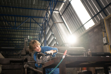 Fototapeta na wymiar Low angle view selective focus of a Caucasian female lathe worker in a blue uniform and gloves, standing at a table with tools, working with a flat iron rod on a bench vise in a metal industry factory