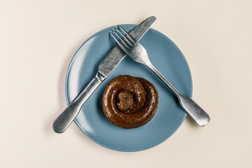 Plastic excrement lies on a plate between a fork and a knife close-up. The concept of an inept cook, fake news, shitty restaurant, bad food, taste of shit