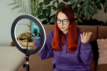 Modern blogger woman in purple hoodie records video on her phone by light of ring lamp at home on...