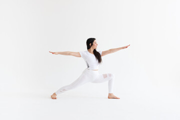 a beautiful young girl with dark hair stands in the pose of Virabhadrasana 2 on a white background. Yoga class