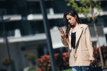 Serious confident female entrepreneur dressed in stylish suit using mobile phone while standing  on street