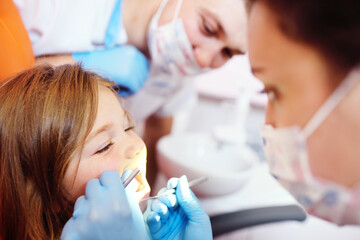 young dentists, a man and a woman, examine the teeth of a child's patient - a little pretty girl who is sitting in an orange dental chair.