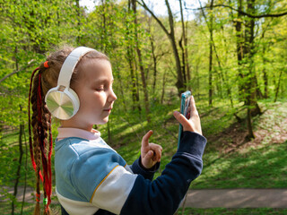 Adorable child girl in headphones learning language listening to music audio books podcasts smartphone outdoor. Blogger kid with gadget distance school online lesson chat cell phone conference in park