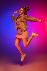 Portrait of young girl in casual cloth jumping, posing isolated over gradient pink blue background in neon light