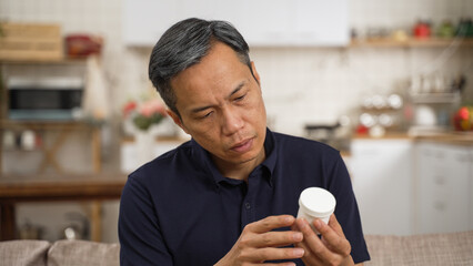 confused asian senior man with bad memory looking at a white container in hand and can’t remember...