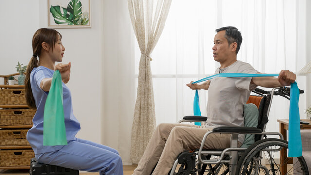 asian woman physiotherapist teaching handicapped older man how to use elastic band while she is doing resistance training on wheelchair at home