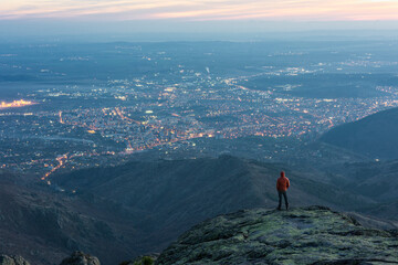 Close up tourist, climber with dark red jacket standing on the top of Balkan mountain,staring at city Sliven at sunset. The shining lights of the city are sparkling in the far distance. Sport, travel