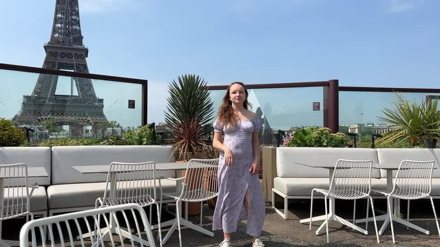 a beautiful teenager girl in a long blue dress stands against the backdrop of the Eiffel Tower in a restaurant, she straightens her hair and leaves the dress is blown by the wind of space for text.