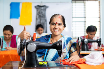 Happy smiling woman showing thumbs up sign by looking camera while working at garments sewing...