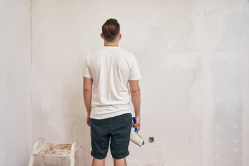 a young man stands sadly and looks at an unpainted wall and holds a roller in his hands. view from the back. renovation concept, home renovation, wall painting