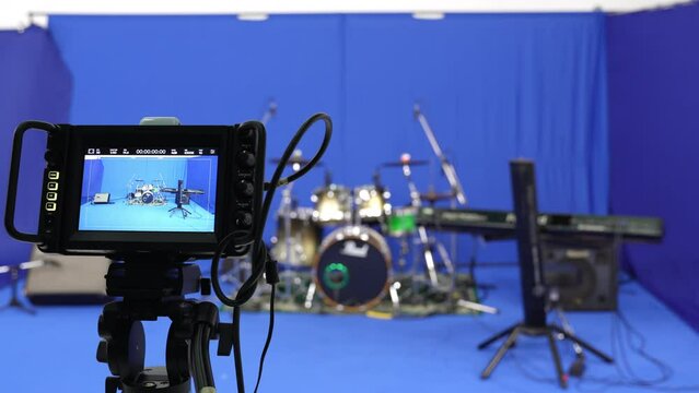 Selective focus and close-up of the video camera screen that capturing instruments with a drum kit, keyboard, and stereo, arranged on the blue screen backdrop in the studio, preparing for filming.
