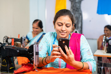 Woman tailor using mobile phone in front of sewing machine at garments - concept of relaxation, technology and checking online designs.