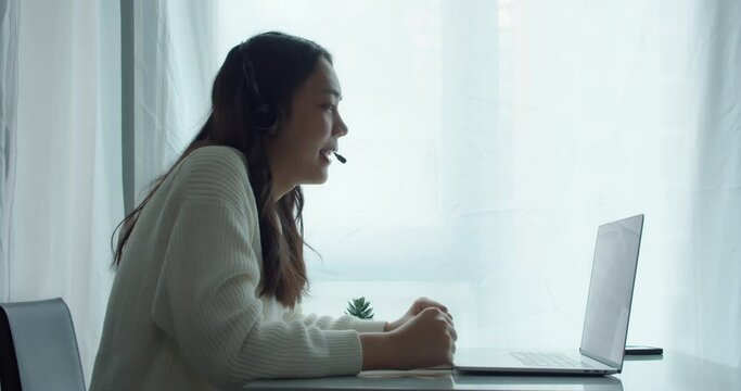 Close up of beautiful asian businesswoman wearing headphones for a videoference from home office.Female are student by webcam video call chat explain and take note course online at home.