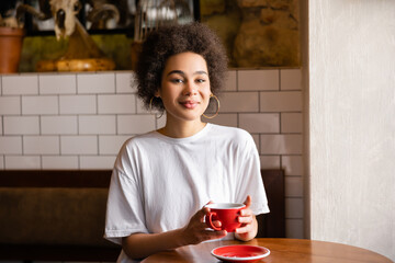 cheerful african american woman in hoop earrings and white t-shirt holding cup