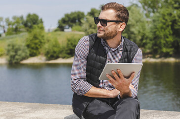 Fototapeta premium Mid adult man in sunglasses holds digital tablet in hand while sits on the waterfront.