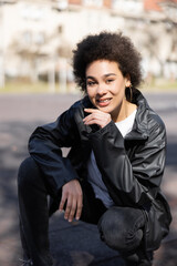 cheerful african american woman in jacket sitting on asphalt