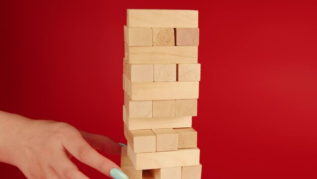 Jenga game on red background. Woman's hand takes out small wooden blocks and they fall. Game over. Unrecognizable person playing board game Jenga. Concept of leisure activity at home.