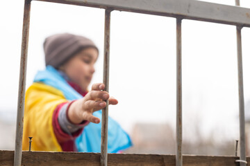 A little refugee girl with a sad look behind a metal fence. Social problem of refugees and internally displaced persons. Russia's war against the Ukrainian people