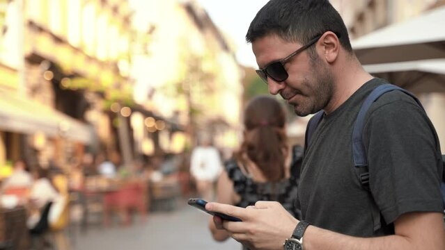 Handsome man in casual clothes and sunglasses walks along the city center street looks around , old city on background. Bearded caucasian traveler uses smartphone and takes pictures. 4k