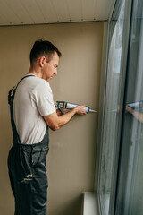 Fototapeta premium Young man wearing overalls sealing cracks between window and trim