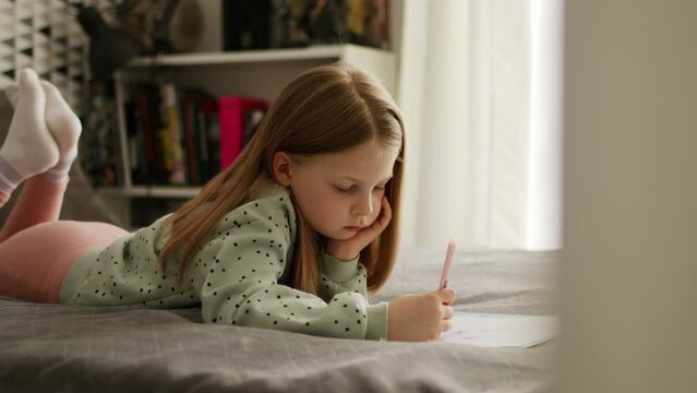 Little kid girl draw or hand writing in diary by pencil. Child learning and doing homework studying at home lying on a bed.	
