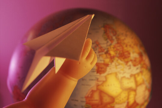 Close-up Of A Doll's Hand Holding A Paper Plane Near A Globe