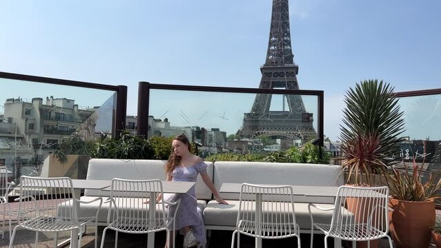 a fair-haired girl in a long beautiful dress sits against the background of the Eiffel Tower with a place for text in a restaurant overlooking the Eiffel Tower She leans back on the seat.