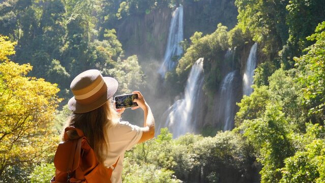 Back view of travel blogger woman on trip or adventure on Tee Lor Su waterfall in Thailand. Young female traveler make photos on smart phone for social media. Generation Z new concept travels