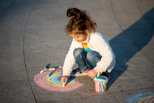 A cute girl draws a rainbow with crayons on the street. Creation. Kids. Street.