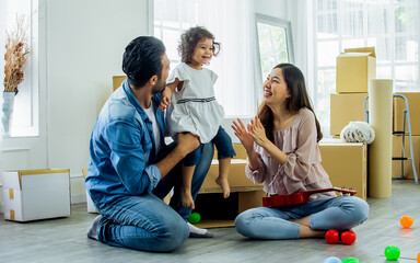 Happy family with three members Caucasian father, Asian mother and little daughter laughing, playing together to hug and lift the girl to the air with fun and happiness while moving to new house.