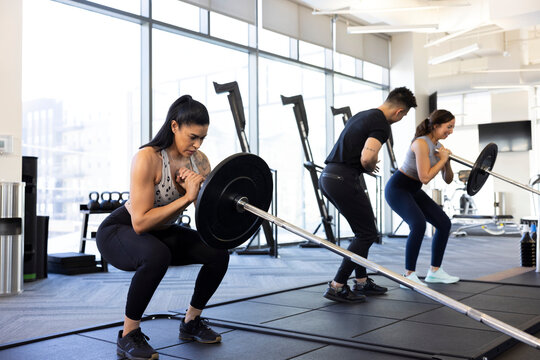 Strength trainer looking on as two woman do squats with barbell.