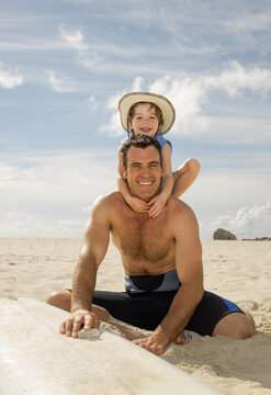 Father smiling and waxing his surfboard while little daughter climbs on his back