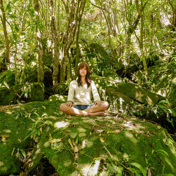 Young woman sitting on a moss covered rock in the lotus position in natural forest