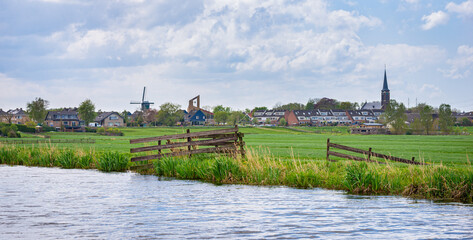 Fototapeta na wymiar Panoramic scenery of the village Hazerswoude-dorp amidst green meadows with wooden fence in the foreground.