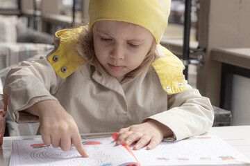 A child sits at the table in a café or terrace outdoors and follows the lines with her fingers in...