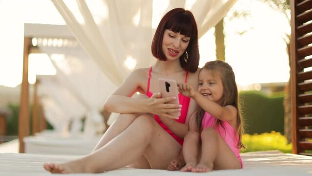 Happy smiling mom holding smartphone with her little kid girl, while resting in the outdoor gazebo near swimming pool having a video call touching the display. Vacation gadgets and technology concept
