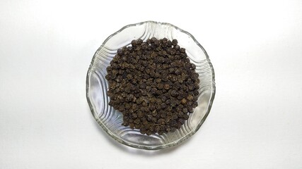 Bunch of organic and farm fresh dried south Indian Kerala spice black pepper seeds inside a transparent glass bowl on a white table surface with copy space. Horizontal closeup macro flat lay top view.