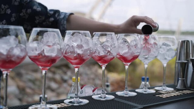 Bartender pours one part of red drink from metal jigger into glasses with ice cubes in row on bar counter in open air. Man in suit walks by, shaker and jigger on bar.