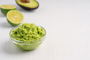 Mexican guacamole vegetarian healthy dip, spread, or salad made of mashed ripe green avocado served in glass bowl on plate with  nachos as snack on white wooden table with lime. Image with copy space